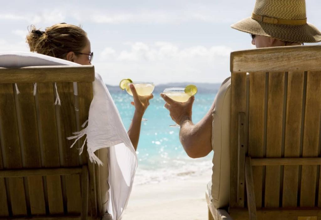 people sit in two wooden beach chairs facing the Caribbean ocean holding drinks and making a toast